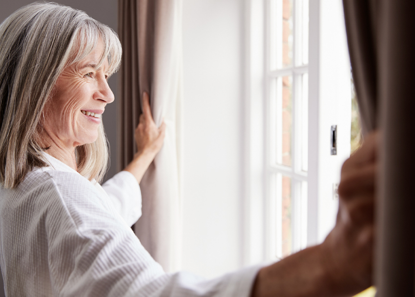 Older woman joyfully opens a living-room window, letting fresh spring air flow inside.