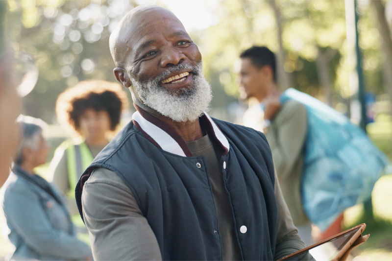 Smiling senior man volunteering at a community event, standing outdoors with other volunteers in the background.