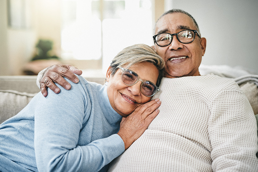 A smiling older couple relaxing on a couch, with the woman leaning comfortably against the man as they embrace in a warm, cozy living room