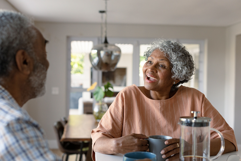 Older adults enjoying coffee together at a kitchen table, smiling and talking in a bright, comfortable home.