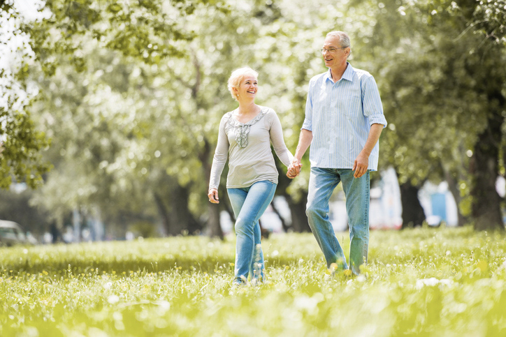 Older couple holding hands and walking through a sunny green park in spring.