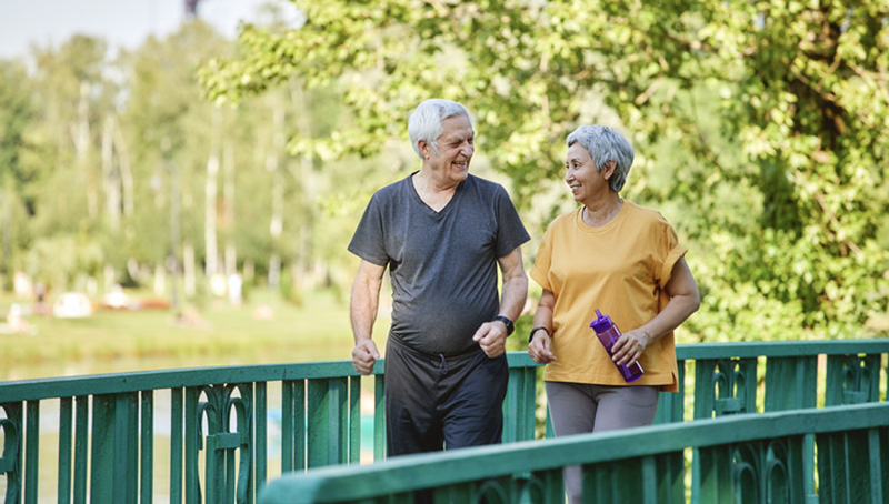 Older couple smiling while walking together across a bridge in a sunny park.